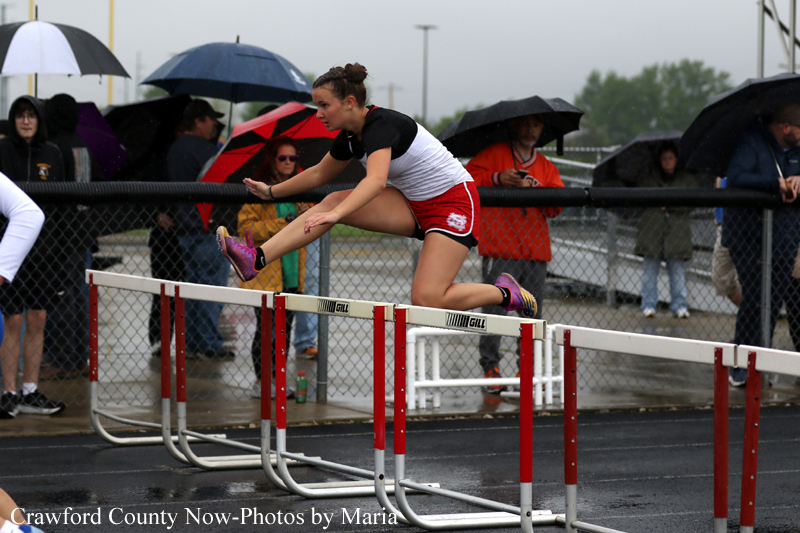 Female hurdler in red shorts and white top clears a hurdle during a rainy track meet, spectators with umbrellas behind a fence.