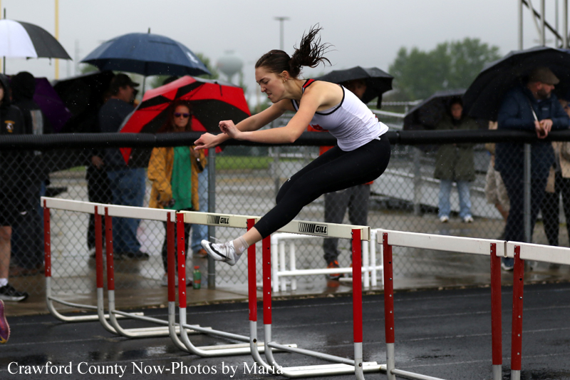Female hurdler in a white tank top and black leggings clears a hurdle on a rainy track while spectators with umbrellas watch.