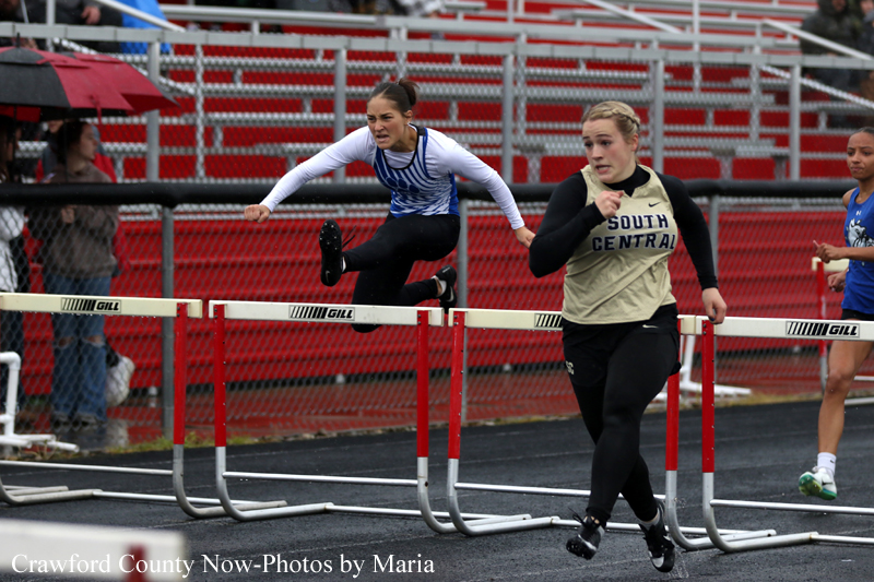 Two female athletes compete in a hurdle race; one clears a hurdle while the other runs beside on a track with red bleachers in the background.