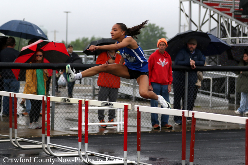 Female hurdler in a blue uniform clear a hurdle on a rain-soaked track while spectators under umbrellas watch in the background.