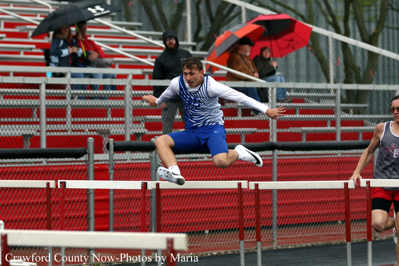 Male high jumper in blue shorts clears the bar during a track meet as spectators with umbrellas watch from red bleachers.