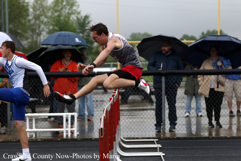 Male hurdler clears a red hurdle on a rainy track as spectators with umbrellas watch in the background.