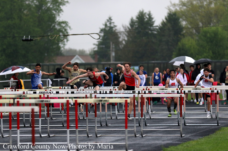Runners jump over hurdles in a crowded track race on a wet, overcast day with spectators and umbrellas in the background.
