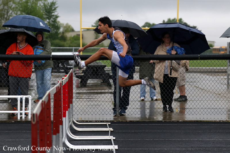 Male hurdler in blue and white uniform clears a hurdle on a rainy track as spectators with umbrellas watch from behind a chain-link fence on a wet athletic field.