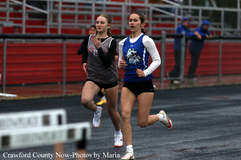 Two female sprinters race on an outdoor track, one in gray/black and the other in blue/white with a lion logo, with stands in the background.