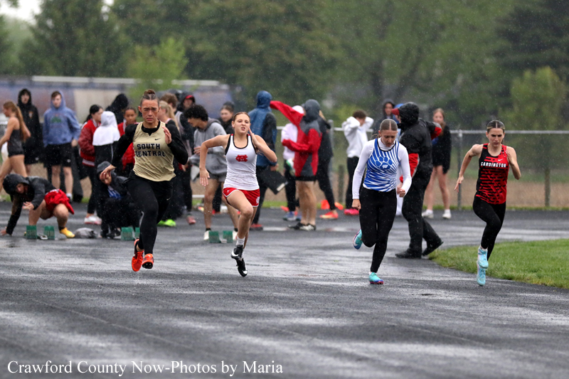 Four young athletes sprint from the starting line on a wet track, with onlookers in the background on a rainy day.