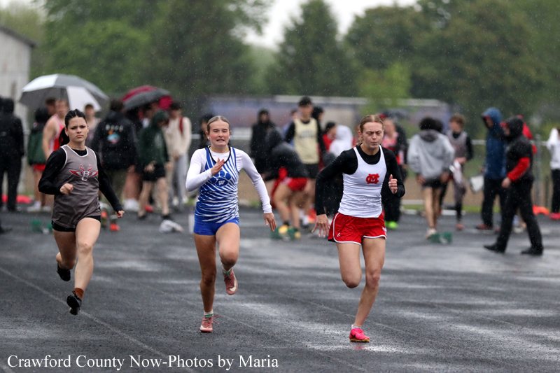 Three female sprinters race on a wet track in the rain, spectators with umbrellas behind them.