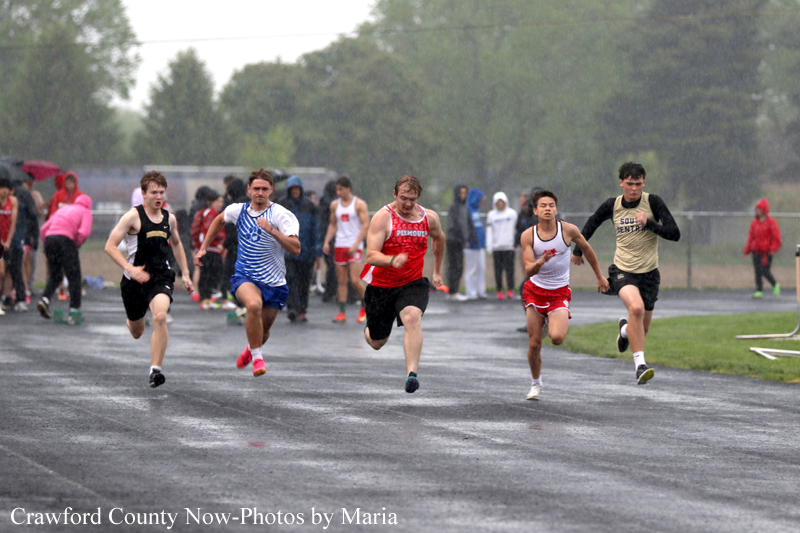 Five male sprinters race on a wet track during a rainy day with spectators in the background.