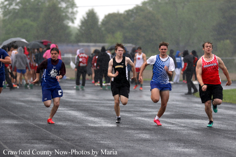 Group of male sprinters racing on a wet track in the rain, with spectators and umbrellas in the background.