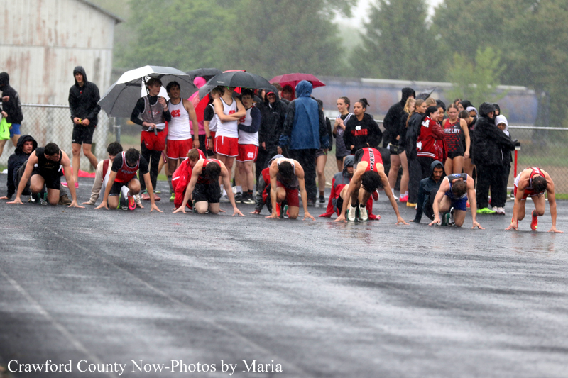 Runners crouch at the starting line on a wet track in the rain, while spectators hold umbrellas in the background.