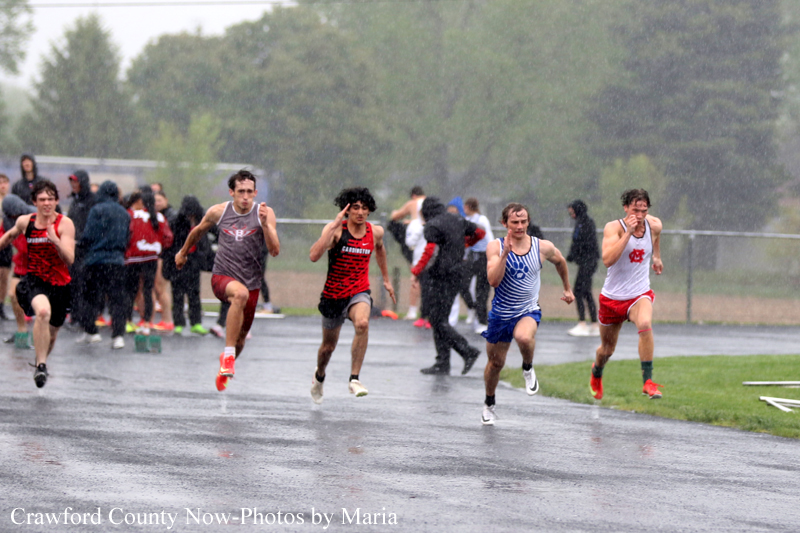 Runners sprinting on a wet outdoor track in light rain, with several competitors in colorful uniforms and spectators in the background.