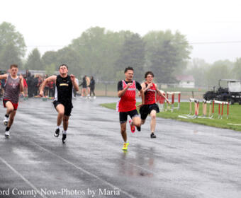 Runners sprint on a wet track during a heavy rain outdoor race, athletes in red and dark uniforms with spectators in the background.