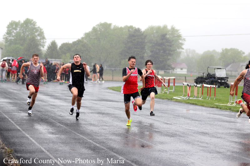 Runners sprint on a wet track during a heavy rain outdoor race, athletes in red and dark uniforms with spectators in the background.