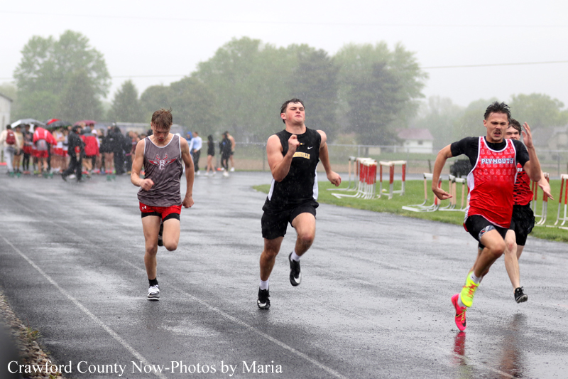 Three male sprinters race on a wet track in the rain, with spectators in the background under umbrellas.
