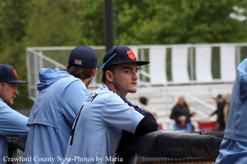 Baseball players in light-blue uniforms lean on a dugout railing; one faces the camera wearing a navy cap with an orange paw logo.
