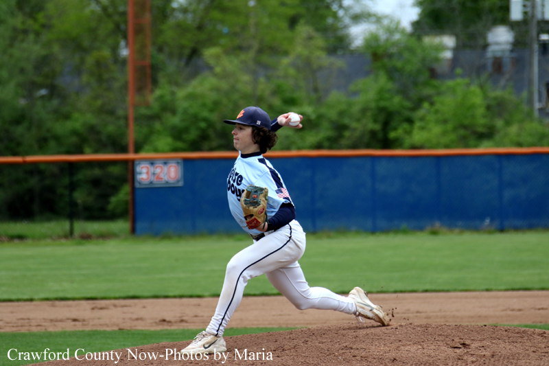 Baseball pitcher in white and blue uniform mid-throw on the mound with a green field and blue fence in the background.