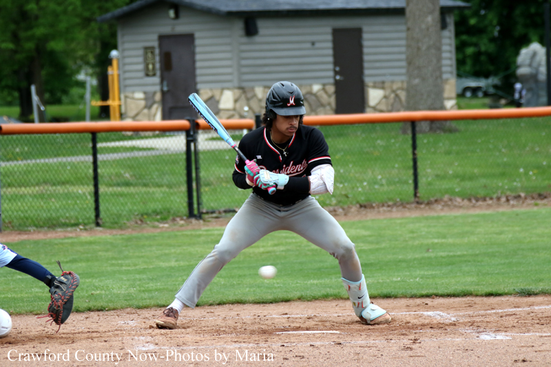 Baseball batter in black uniform and helmet prepares to swing as a pitched ball approaches on a dirt infield.