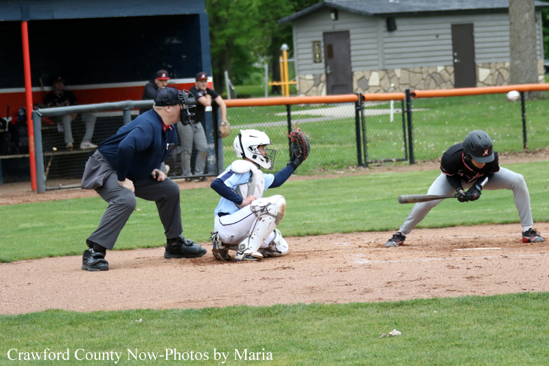 Baseball action at home plate: catcher in white gear crouches as an umpire watches, while a batter/runner approaches the plate with a bat nearby.