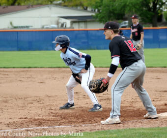 Youth baseball game scene: a baserunner in a light blue uniform slides toward a base as a fielder in a black jersey (#12) waits with a glove nearby on the dirt infield.