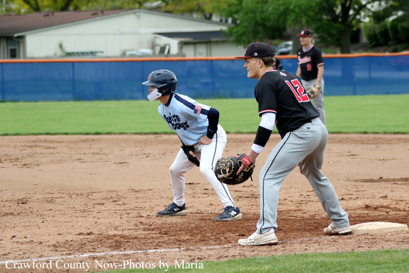 Youth baseball game scene: a baserunner in a light blue uniform slides toward a base as a fielder in a black jersey (#12) waits with a glove nearby on the dirt infield.