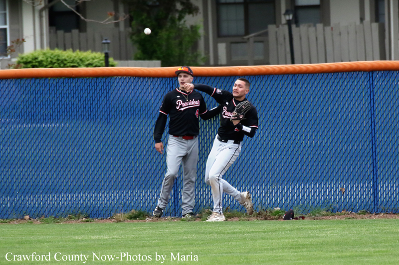 Two baseball players in black uniforms with pink lettering stand near a blue chain-link outfield fence as a ball flies between them during a play.