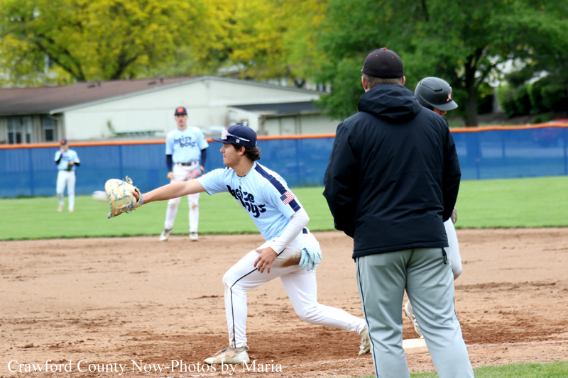 Young baseball pitcher in a light blue jersey and white pants lunges to throw, glove outstretched, as a coach in a black jacket watches nearby on the dirt infield.