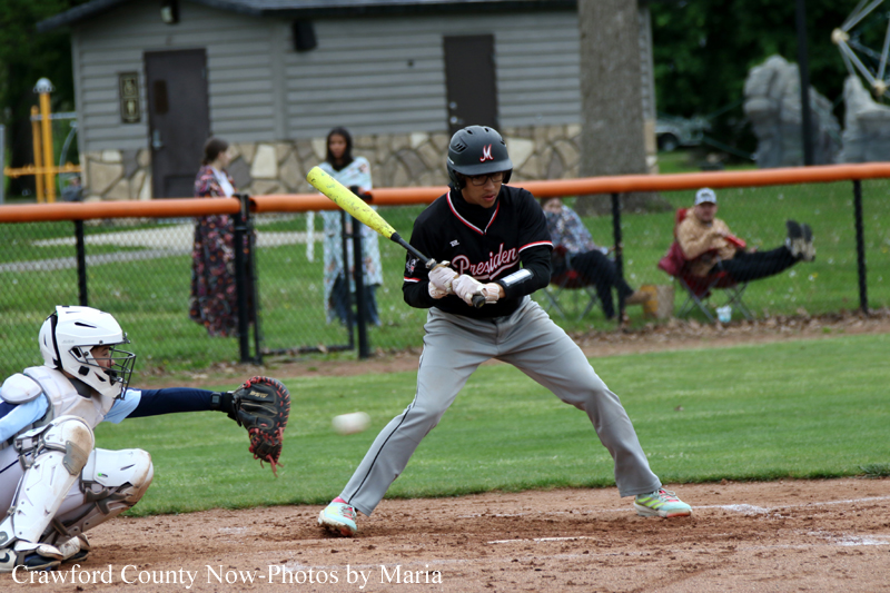 Baseball batter in a black jersey swinging at a pitch, with a catcher in white gear kneeling behind him on the dirt infield.