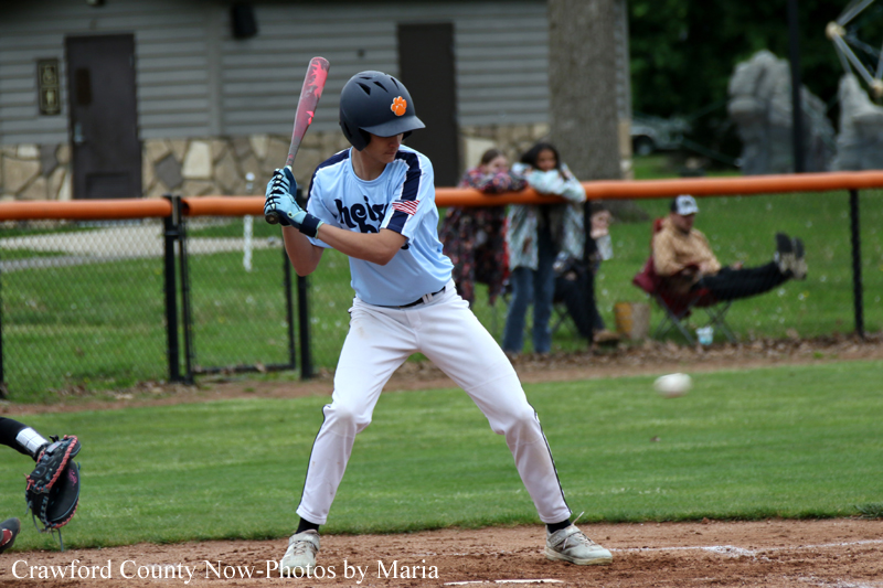Young baseball batter in a light blue jersey and white pants stands at the plate, preparing to swing as a catcher looms to the left and spectators watch in the background.