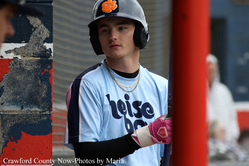 Young baseball player in a light blue jersey and black sleeves stands with a bat, wearing a helmet with an orange paw logo and pink batting gloves beside a red post.
