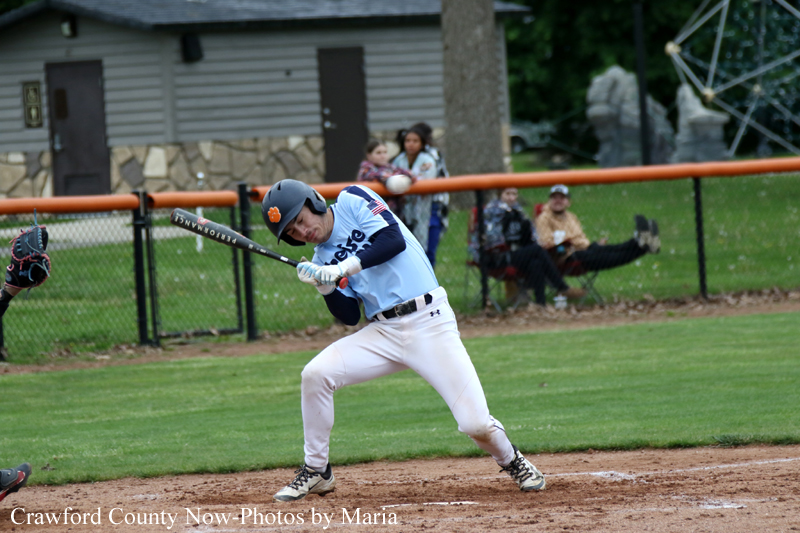 Baseball batter in light blue uniform swings at a pitch on a dirt infield with a catcher visible to the left and spectators behind the fence.