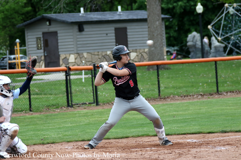 Male baseball batter in a black 'Presidents' jersey and gray pants grips the bat, ready to swing at home plate as a catcher in white gear crouches behind him.