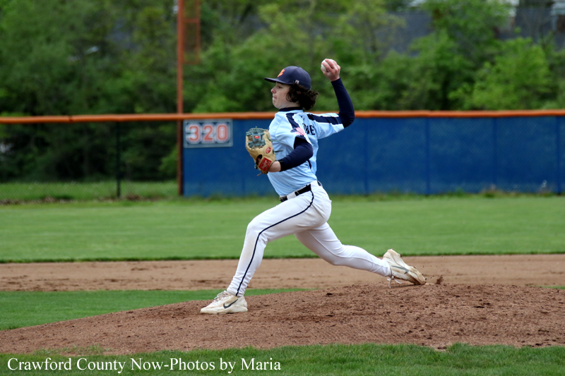Young baseball pitcher in white uniform on the mound, mid-throw with glove up, action shot on a grassy field with a blue outfield wall in the background