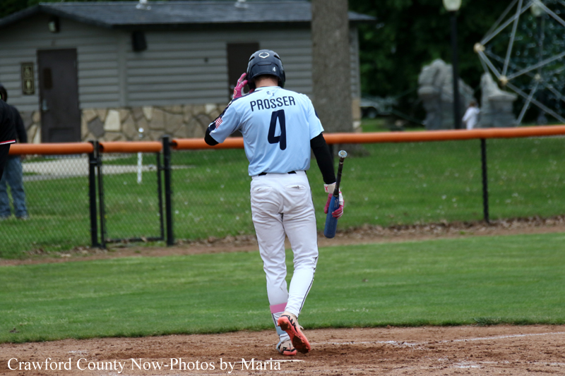 Baseball player in light blue jersey with 'Prosser' and number 4 walking on infield with helmet and bat in hand (rear view).