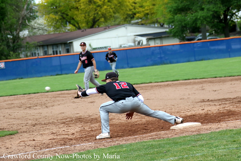 Baseball game action: a fielder in black number 12 lunges toward second base as players watch in the infield with a green outfield and blue fence in the background.
