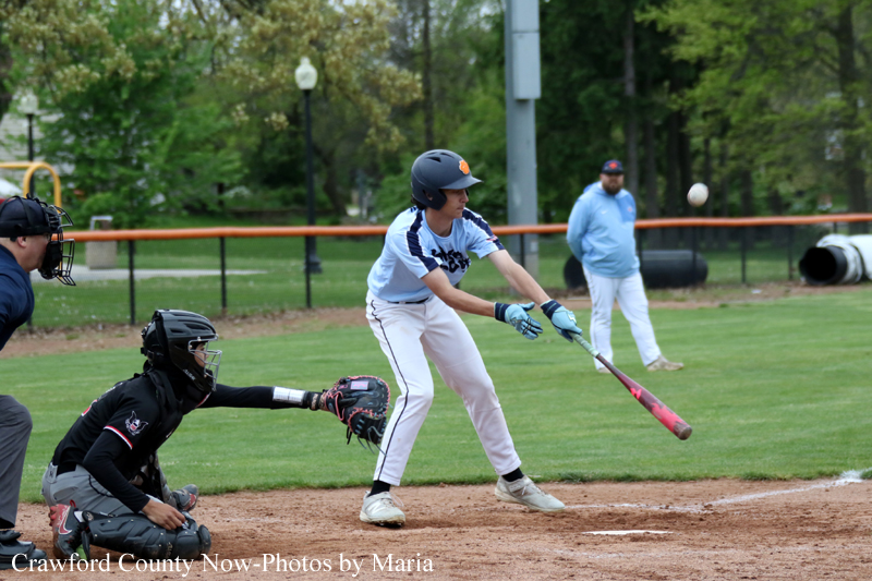 Baseball batter in light blue uniform swings at a pitched ball as the catcher in black crouches behind him and an umpire watches; ball is midair to the right on a grassy field.