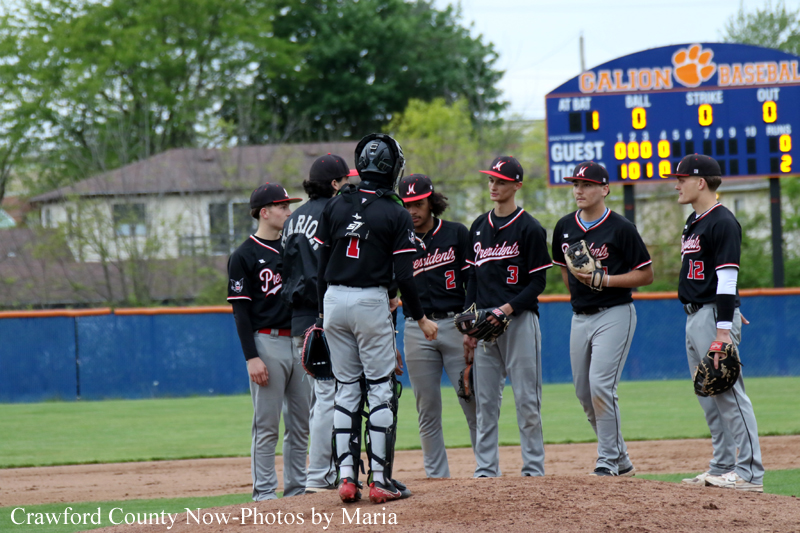 Young baseball team in black-and-red uniforms gathers on the pitcher's mound, a catcher addressing players as a scoreboard looms in the background.