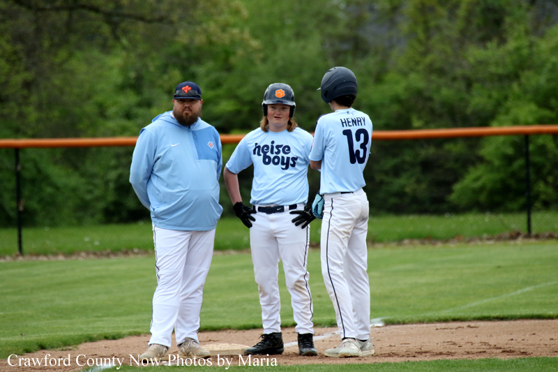 Three baseball players stand on a dirt infield with a coach on a green field, wearing light blue uniforms; one has hands on hips and wears a helmet, the others chat nearby.
