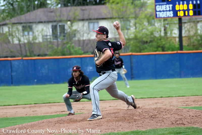 Baseball pitcher in black uniform mid-pitch on the mound with catcher behind him and a scoreboard in the background.