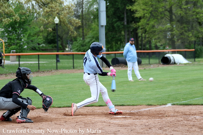 Young baseball batter in mid-swing, catcher crouched behind him, coach or umpire in background on a grassy field.