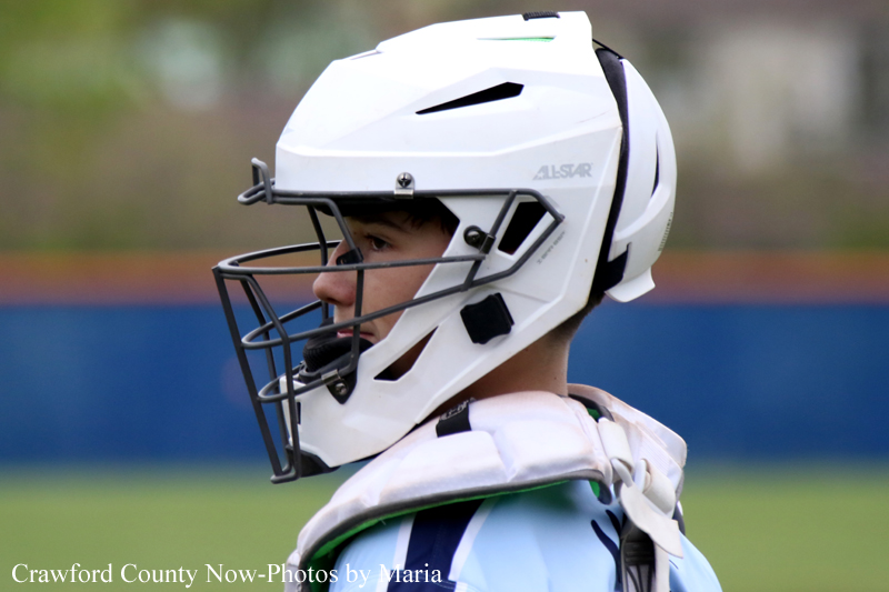 Side profile of a lacrosse goalie wearing a white protective helmet with face mask on a field.
