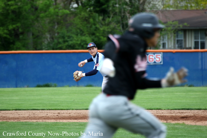 Baseball game in progress: a fielder in blue-and-white uniform prepares to catch the ball, while a blurred runner or fielder passes in the foreground on the green field.