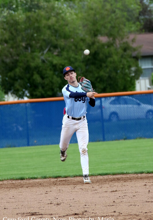 Baseball pitcher in a light blue uniform throwing a pitch on a dirt mound, ball in midair with a blue outfield fence behind.