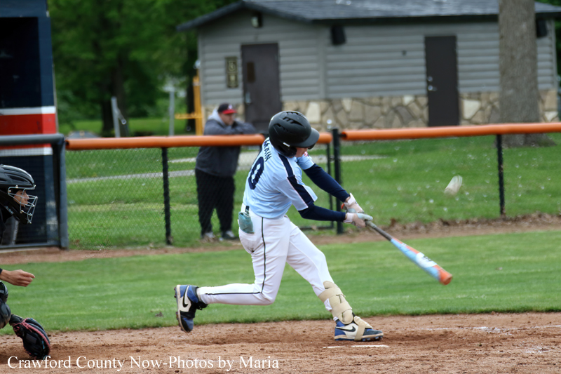 Youth baseball batter in white and blue swings at a pitched ball on a dirt infield, with a catcher in black gear nearby.