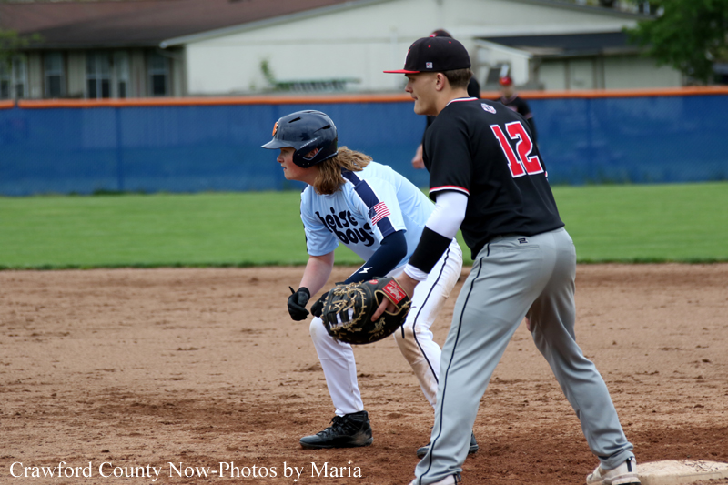 Baseball scene: a fielder in a light-blue uniform crouches with a glove as a runner in a black uniform with the number 12 stands nearby on the dirt infield.