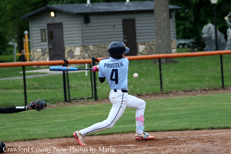 Baseball batter in a white uniform swings at a pitched ball on a diamond with a catcher nearby and a fence in the background.