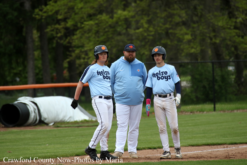 Three baseball players in light-blue uniforms with helmets pose with their coach on the infield dirt.