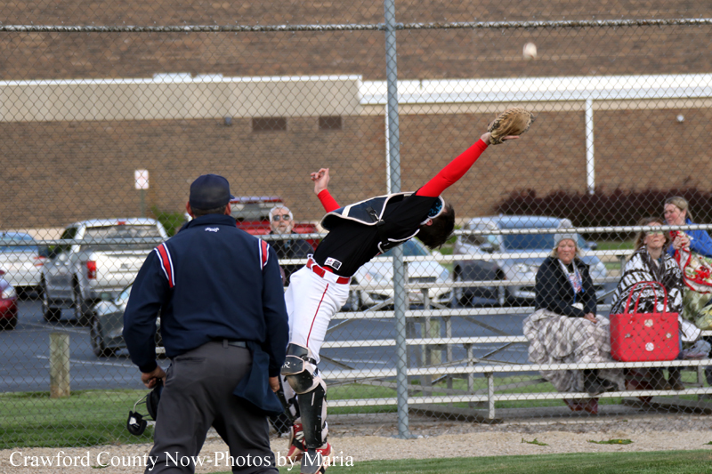 Baseball scene: catcher in black gear and red sleeves flipping mid-air near home plate amid an umpire and spectators behind a chain-link fence.