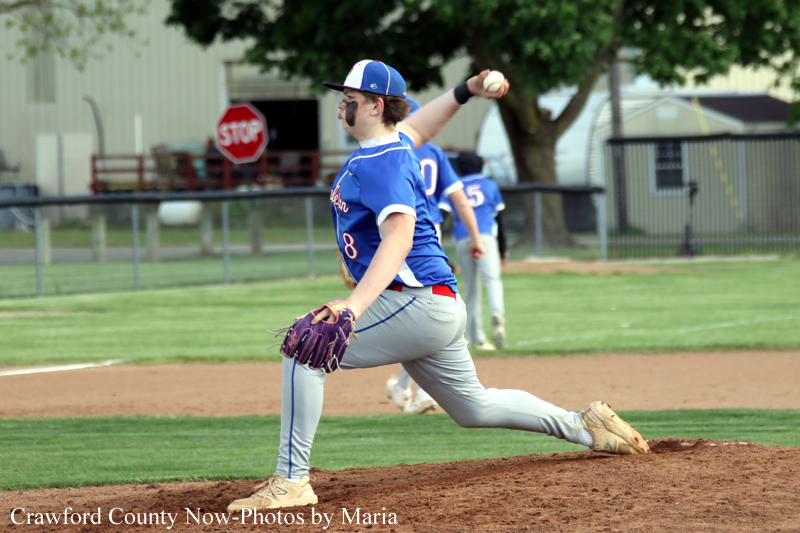 A baseball pitcher in a blue uniform winds up on the mound, throwing toward home plate with a glove on his left hand and a teammate visible in the background.