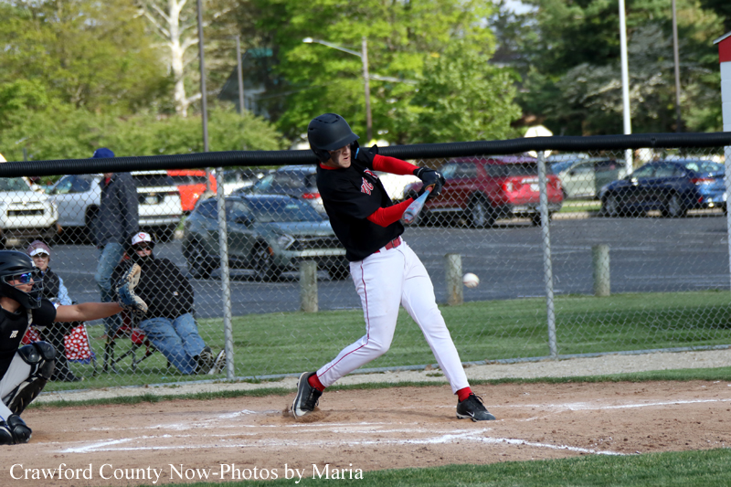 Baseball batter in black and red, mid-swing at home plate with catcher nearby and spectators behind a chain-link fence.