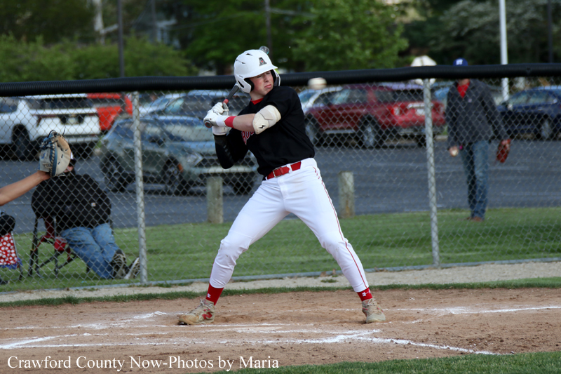 Young baseball batter in a white helmet and white pants swings at a pitch from the plate during a game, with a chain-link fence in the background.